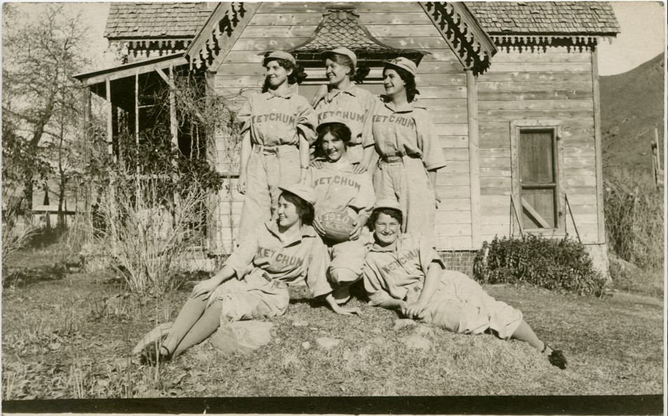 Six women pose in baseball uniforms in front of a house in 1910.