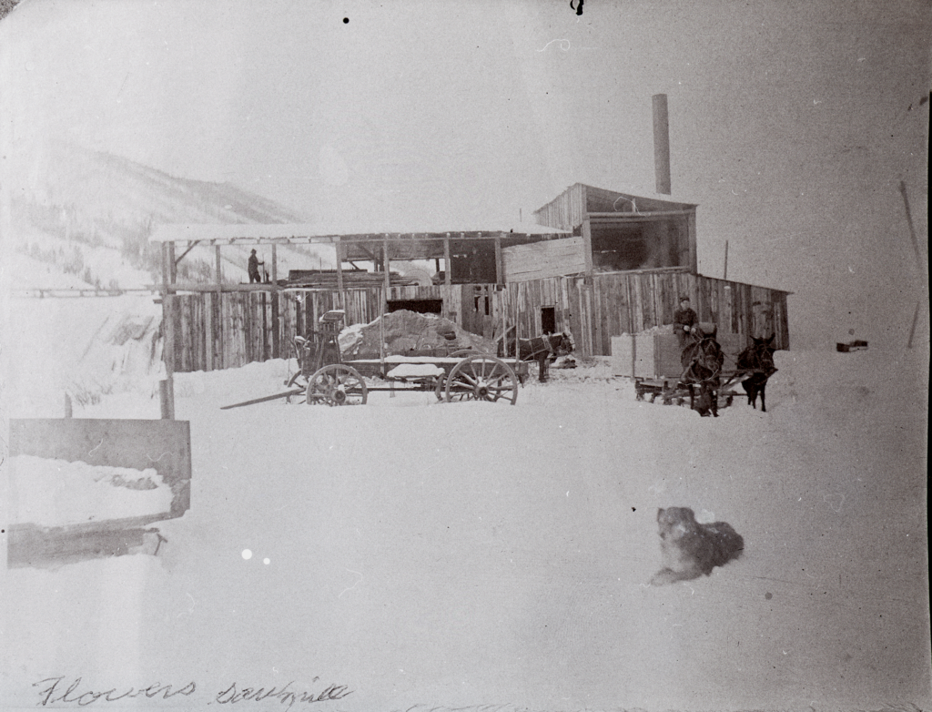 A long wooden building in a snowy mountain-scape, with workers, a wagon, and a sleigh in front.