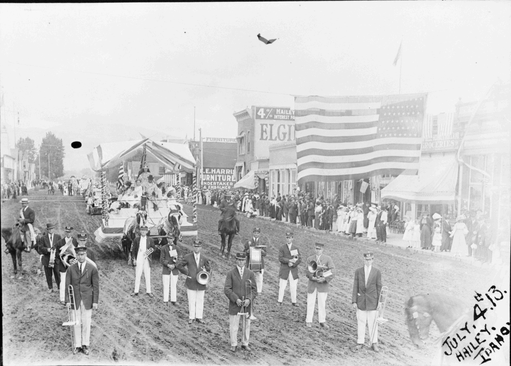Men stand on a dirt street with a large American flag and a parade float.